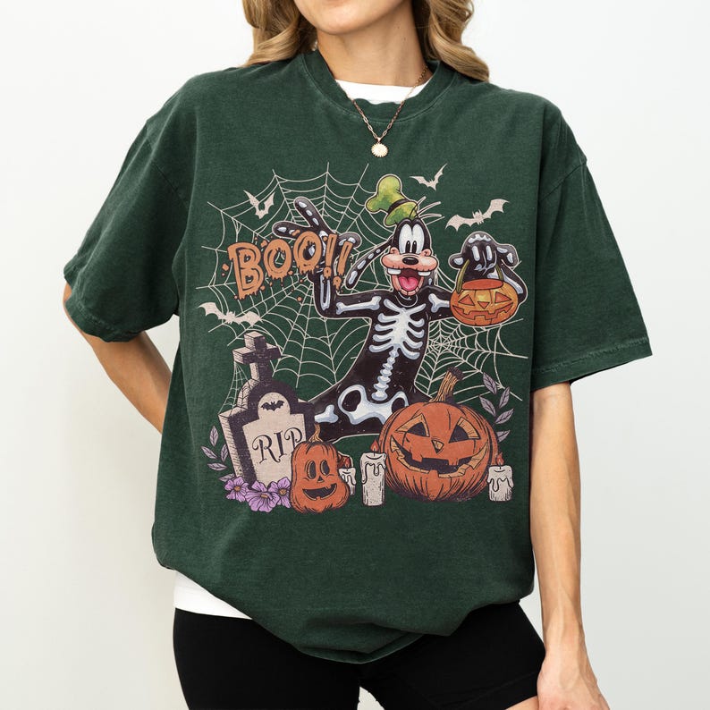 May include: A dark green short-sleeve t-shirt featuring a graphic of a cartoon dog in a skeleton costume, a tombstone with the text RIP, a jack-o'-lantern, and the word BOO. The design includes a spiderweb background with bats and candles.