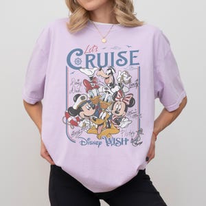 May include: A light purple t-shirt featuring a graphic of Disney characters, including Mickey Mouse, Minnie Mouse, Donald Duck, and Goofy. The design includes the text "Let's Cruise" and "Disney Wish" in a nautical style. The shirt has short sleeves.