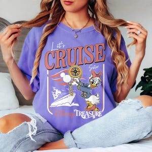 May include: A purple t-shirt with the words "Let's Cruise Sister" in a retro font. The graphic features Daisy Duck holding a compass, a cruise ship, and the words "Disney Treasure".