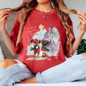 Pu&ograve; includere: T-shirt rossa con un'illustrazione di Topolino e Minnie in abiti natalizi, un castello sullo sfondo e la scritta "It's the most magical time of the year."