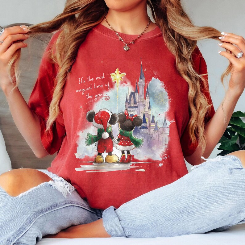Pu&ograve; includere: T-shirt rossa con un'illustrazione di Topolino e Minnie in abiti natalizi, un castello sullo sfondo e la scritta "It's the most magical time of the year."