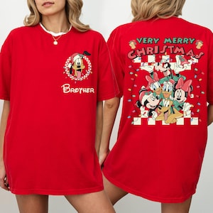 May include: Red t-shirt featuring a festive design. The front displays a cartoon character in a Santa hat with the word "Brother". The back showcases a group of cartoon characters with the text "Very Merry Christmas" and string lights.
