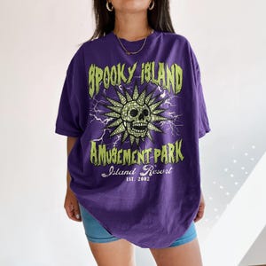 K&ouml;nnte beinhalten: Ein lila T-Shirt mit einem Aufdruck mit der Aufschrift "Spooky Island Amusement Park Island Resort Est. 2002". Die Grafik zeigt einen Totenkopf mit Sonnenstrahlen und Blitzen. Das Shirt ist &uuml;bergro&szlig;.