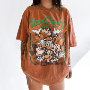 May include: An orange short-sleeve t-shirt featuring a graphic of Disney characters in skeleton costumes. The text reads Mickey's Not-So-Scary Halloween Party and Walt Disney World. The design includes Mickey, Minnie, Donald, Daisy, and Goofy.