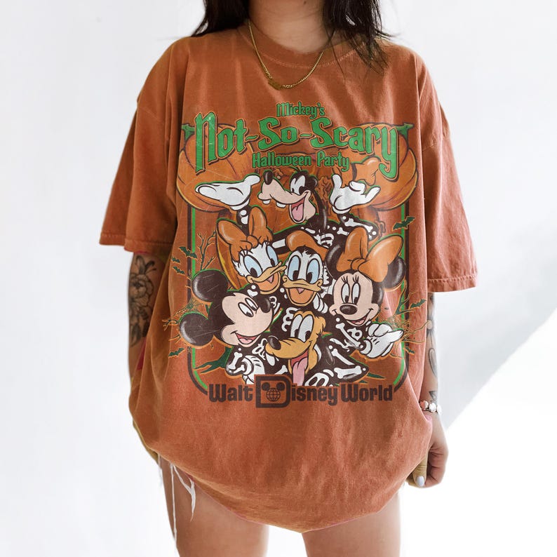 May include: An orange short-sleeve t-shirt featuring a graphic of Disney characters in skeleton costumes. The text reads Mickey's Not-So-Scary Halloween Party and Walt Disney World. The design includes Mickey, Minnie, Donald, Daisy, and Goofy.