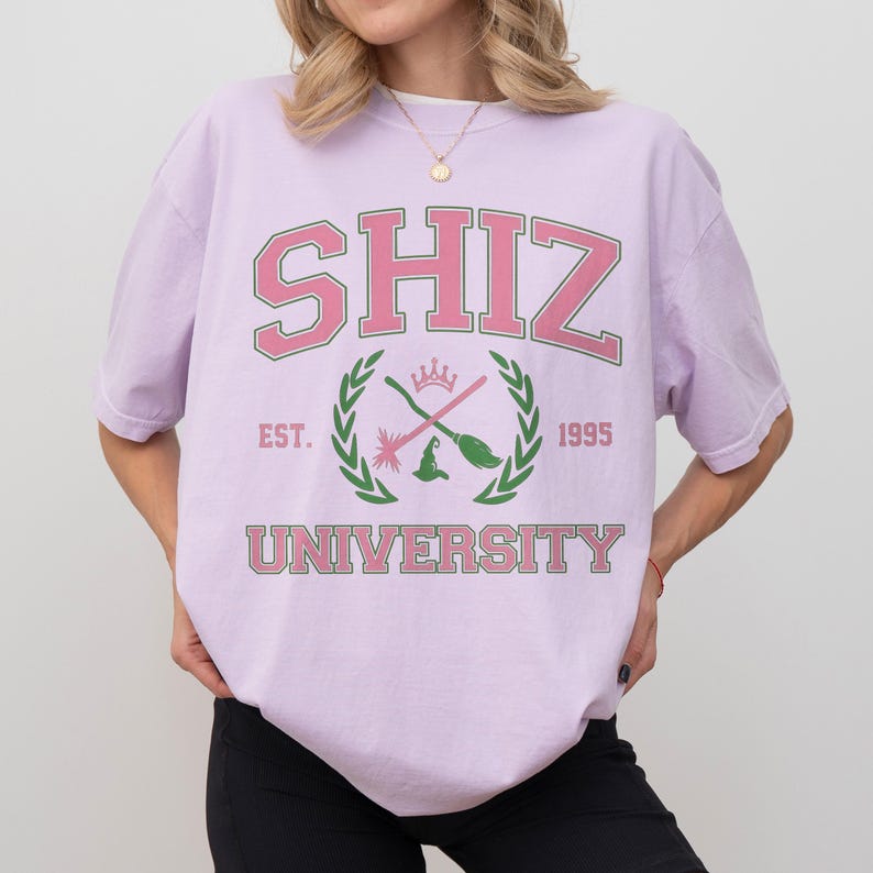 Pu&ograve; includere: Una maglietta lilla con la scritta "SHIZ UNIVERSITY" in rosa e verde. Il design include uno stemma con un cappello da strega, una scopa e una corona. La maglietta ha anche la scritta "EST. 1995".