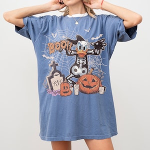 May include: A blue short-sleeved t-shirt featuring a graphic print of a cartoon duck in a skeleton costume, a tombstone with the text RIP, pumpkins, and spiderwebs. The design includes the word BOO in orange lettering against a blue background.
