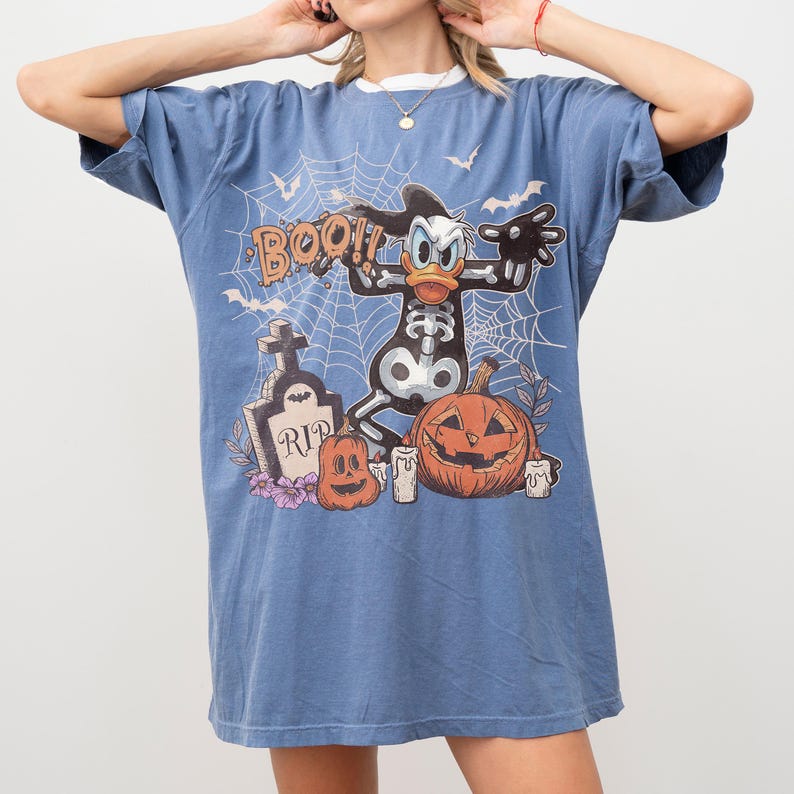 May include: A blue short-sleeved t-shirt featuring a graphic print of a cartoon duck in a skeleton costume, a tombstone with the text RIP, pumpkins, and spiderwebs. The design includes the word BOO in orange lettering against a blue background.