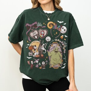 May include: Dark green t-shirt with a colourful graphic design featuring characters from "The Nightmare Before Christmas." The design includes Jack Skellington, Sally, Zero, Oogie Boogie, and other iconic imagery. The shirt has a crew neck and short sleeves.