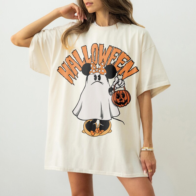 May include: Cream-colored Halloween t-shirt featuring a cartoon ghost with Minnie Mouse ears and a polka-dot bow. The ghost holds a jack-o'-lantern and is set against the word "HALLOWEEN" in orange. A fun, festive design for the spooky season.