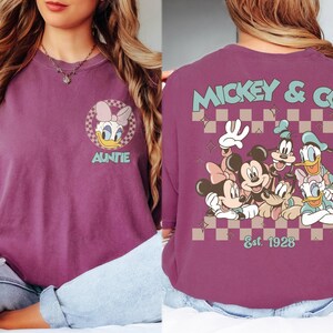 K&ouml;nnte beinhalten: Pflaumenfarbenes T-Shirt mit Daisy Duck und dem Wort "Auntie" sowie einem Mickey & Co.-Design mit Mickey, Minnie, Goofy, Donald und Daisy Duck. Auf der R&uuml;ckseite des Shirts steht "Mickey & Co." und "Est. 1928".