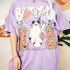 May include: A lilac t-shirt with a Halloween graphic. The design features a ghost, pumpkins, a castle, and the text "Boo To You". A Mickey Mouse head is incorporated into the design.