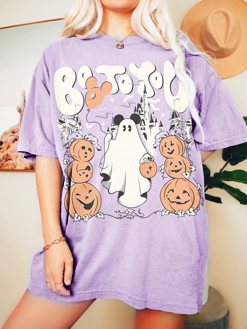 May include: A lilac t-shirt with a Halloween graphic. The design features a ghost, pumpkins, a castle, and the text "Boo To You". A Mickey Mouse head is incorporated into the design.
