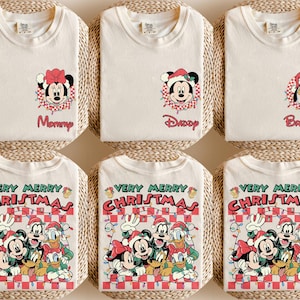 May include: Cream-coloured t-shirts featuring festive graphics. Designs include Minnie Mouse, Mickey Mouse, and Pluto in Santa hats, and a group graphic with the text "VERY MERRY CHRISTMAS". The shirts are displayed on woven mats.