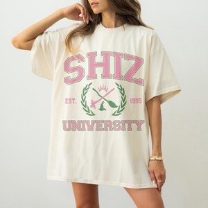 Pu&ograve; includere: T-shirt oversize color crema con la scritta "SHIZ UNIVERSITY" in rosa e verde. Il design include uno stemma con una corona, scope incrociate e l'anno "1995". Una maglietta casual e confortevole.