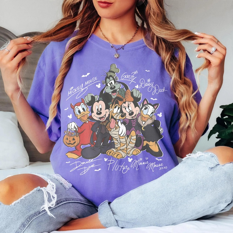 Puede incluir: Una camiseta morada con un estampado gr&aacute;fico de personajes de Disney vestidos con disfraces de Halloween, incluyendo a Mickey Mouse, Minnie Mouse, Donald, Goofy, Pluto y Daisy, con sus nombres escritos en letra blanca alrededor del dibujo.