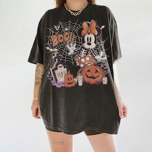 May include: A charcoal black oversized t-shirt featuring a graphic print of a skeleton-themed mouse character, a spider web, a tombstone with the text RIP, pumpkins, and the word BOO. The design includes orange, white, and purple accents.