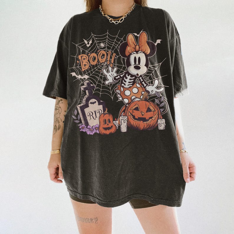 May include: A charcoal black oversized t-shirt featuring a graphic print of a skeleton-themed mouse character, a spider web, a tombstone with the text RIP, pumpkins, and the word BOO. The design includes orange, white, and purple accents.