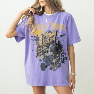 May include: A lilac-colored t-shirt featuring a graphic of Mickey Mouse in a bellhop uniform in front of the Hollywood Tower Hotel. The text "THE TOWER OF TERROR" is printed above the image in yellow. The shirt has short sleeves and a relaxed fit.
