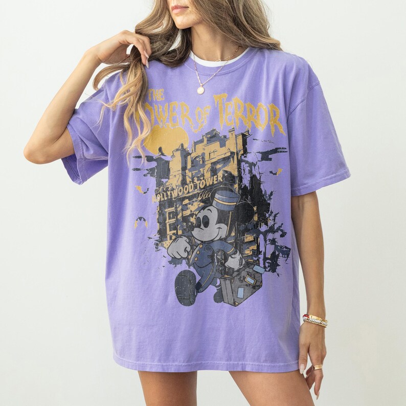May include: A lilac-colored t-shirt featuring a graphic of Mickey Mouse in a bellhop uniform in front of the Hollywood Tower Hotel. The text "THE TOWER OF TERROR" is printed above the image in yellow. The shirt has short sleeves and a relaxed fit.