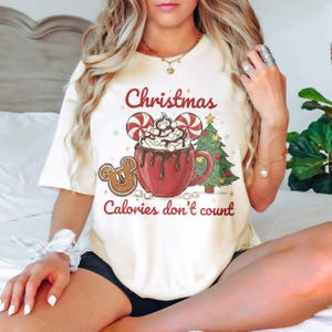 May include: Cream-colored t-shirt featuring a Christmas-themed graphic. The design includes a red mug of hot chocolate with marshmallows, a gingerbread cookie, a Christmas tree, and the text "Christmas Calories don't count."