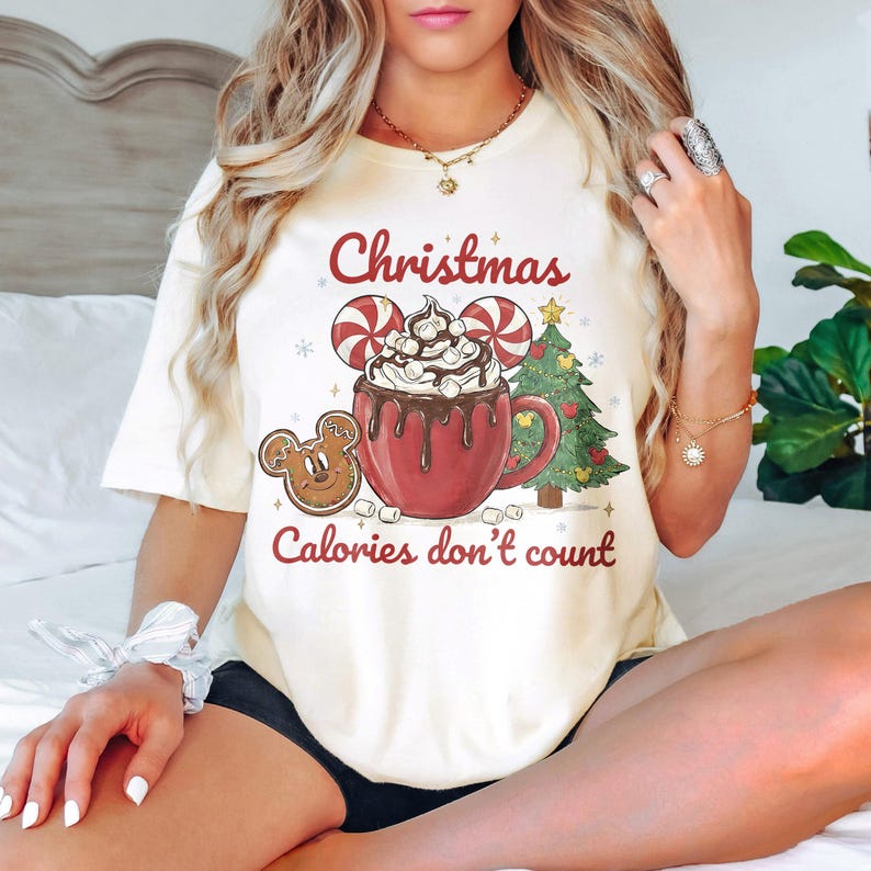 May include: Cream-colored t-shirt featuring a Christmas-themed graphic. The design includes a red mug of hot chocolate with marshmallows, a gingerbread cookie, a Christmas tree, and the text "Christmas Calories don't count."
