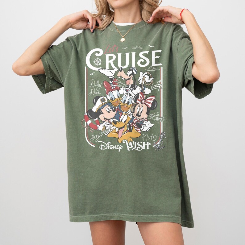 May include: Olive green t-shirt featuring a graphic of Disney characters, including Mickey Mouse, Minnie Mouse, Donald Duck, and Pluto. The shirt has the text "Let's Cruise" and "Disney Wish" in white.