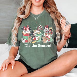 Puede incluir: Camiseta verde con un dise&ntilde;o navide&ntilde;o. Presenta a los personajes de Mickey y Minnie Mouse en tazas festivas, hombres de jengibre y el texto "'Tis the Season!" Una camiseta gr&aacute;fica de temporada para las fiestas.
