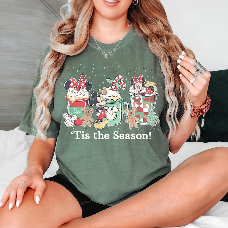Puede incluir: Camiseta verde con un dise&ntilde;o navide&ntilde;o. Presenta a los personajes de Mickey y Minnie Mouse en tazas festivas, hombres de jengibre y el texto "'Tis the Season!" Una camiseta gr&aacute;fica de temporada para las fiestas.