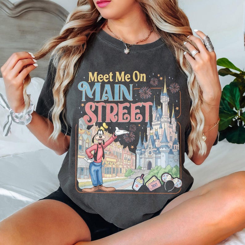 K&ouml;nnte beinhalten: Dunkelgraues T-Shirt mit dem Text "Meet Me On Main Street" in Retro-Schrift. Die Grafik zeigt Goofy, ein Schloss und Feuerwerk. Das Design ist mit einem braunen Rahmen versehen, der eine Vintage-&Auml;sthetik hervorruft.