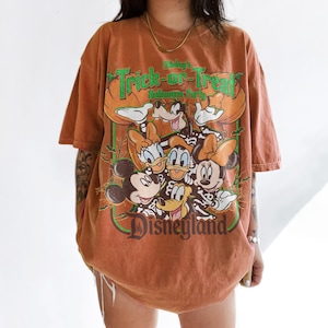 K&ouml;nnte beinhalten: Ein rostfarbenes Kurzarm-T-Shirt mit einem Grafikdruck von Disney-Figuren in Skelettkost&uuml;men. Der Text lautet Mickey's Trick-or-Treat Halloween Party und Disneyland. Das Design zeigt Mickey, Minnie, Donald, Daisy und Goofy.