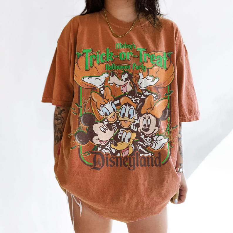 K&ouml;nnte beinhalten: Ein rostfarbenes Kurzarm-T-Shirt mit einem Grafikdruck von Disney-Figuren in Skelettkost&uuml;men. Der Text lautet Mickey's Trick-or-Treat Halloween Party und Disneyland. Das Design zeigt Mickey, Minnie, Donald, Daisy und Goofy.