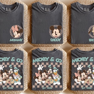 K&ouml;nnte beinhalten: Sechs anthrazitfarbene T-Shirts mit Disney-Charaktergrafiken. Drei Shirts haben individuelle Charakterdesigns mit den Worten "Mommy", "Daddy" und "Brother". Die anderen drei Shirts zeigen eine Gruppengrafik mit "Mickey & Co Est. 1928".