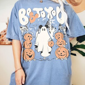 May include: Pale blue t-shirt with a Halloween graphic. The design includes a ghost, pumpkins, and a castle, with the text "Boo to You!" in a whimsical font. The shirt has short sleeves and a loose fit.