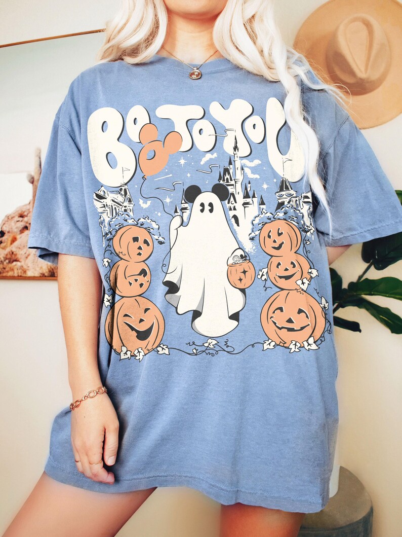 May include: Pale blue t-shirt with a Halloween graphic. The design includes a ghost, pumpkins, and a castle, with the text "Boo to You!" in a whimsical font. The shirt has short sleeves and a loose fit.