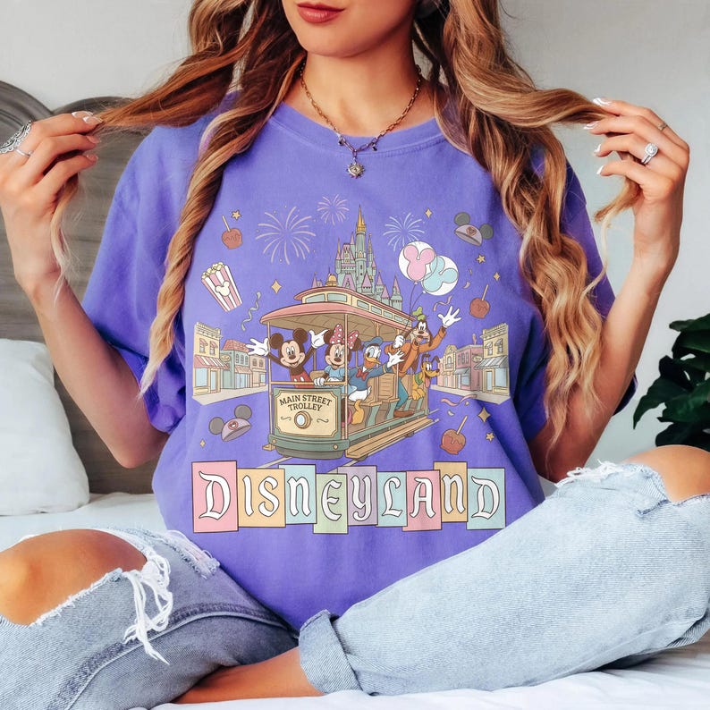 K&ouml;nnte beinhalten: Ein lila T-Shirt mit einem Disneyland-Motiv. Die Grafik zeigt eine Stra&szlig;enbahn mit Mickey, Minnie, Donald und Goofy, sowie Feuerwerk, Popcorn und kandierte &Auml;pfel. Das Wort "Disneyland" ist in Pastellfarben geschrieben.