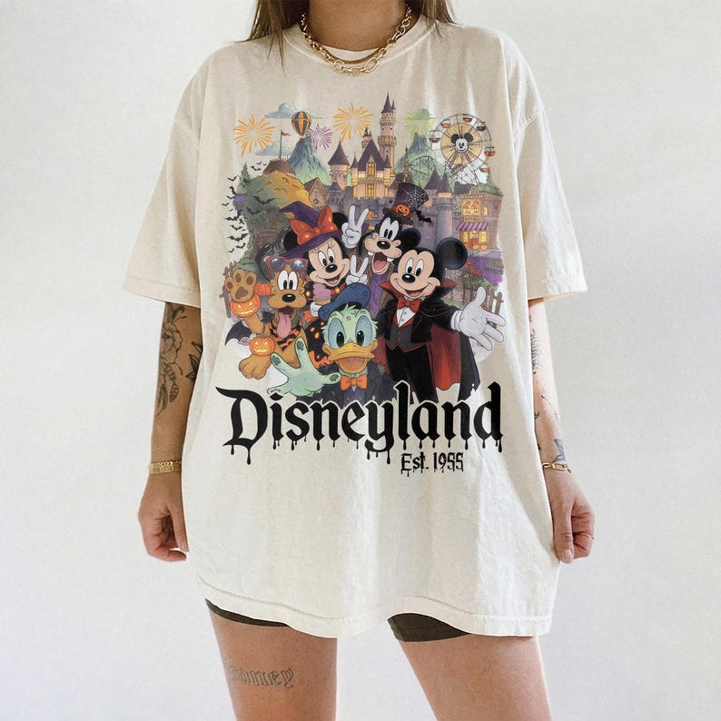 May include: An off-white short-sleeve t-shirt featuring a colorful graphic of Disney characters in Halloween costumes in front of a castle. The text Disneyland Est. 1955 is printed in a black dripping font below the characters. The shirt has a crew neck.