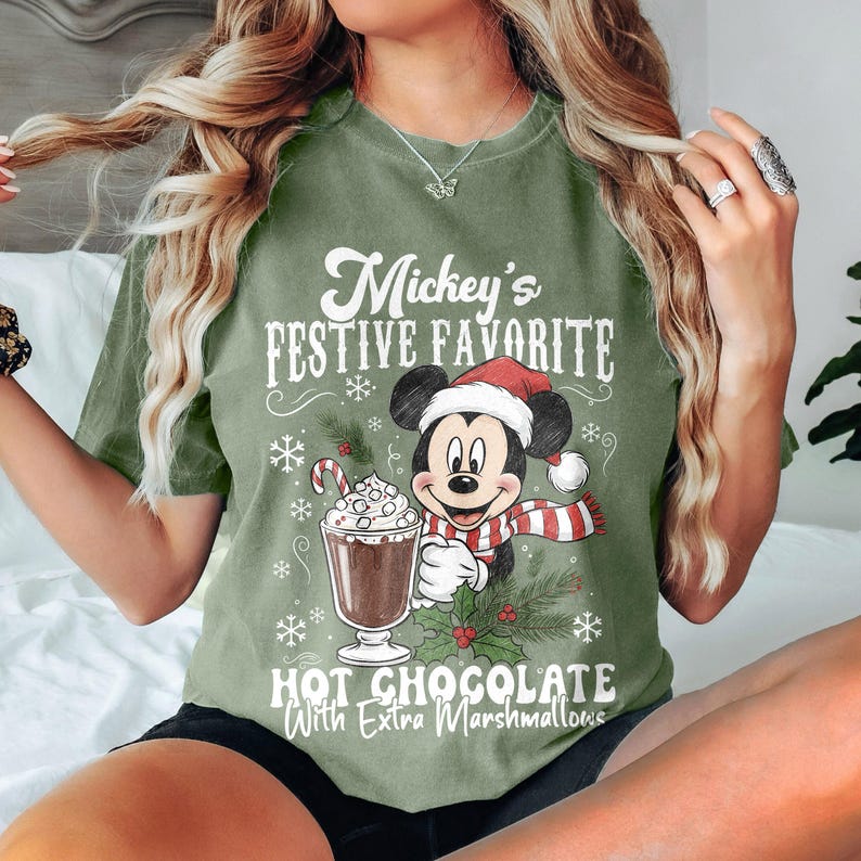 May include: Olive green t-shirt featuring Mickey Mouse in a Santa hat and scarf, holding a hot chocolate with marshmallows. The text reads "Mickey's Festive Favorite" and "Hot Chocolate With Extra Marshmallows".