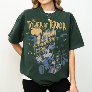 May include: Dark green t-shirt featuring a graphic of Mickey Mouse in a bellhop uniform in front of the Hollywood Tower Hotel. The text "The Tower of Terror" is printed above the image, with "Hollywood Tower Hotel" below.