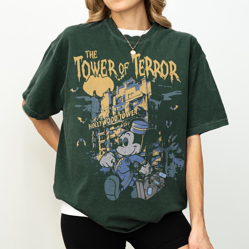 May include: Dark green t-shirt featuring a graphic of Mickey Mouse in a bellhop uniform in front of the Hollywood Tower Hotel. The text "The Tower of Terror" is printed above the image, with "Hollywood Tower Hotel" below.