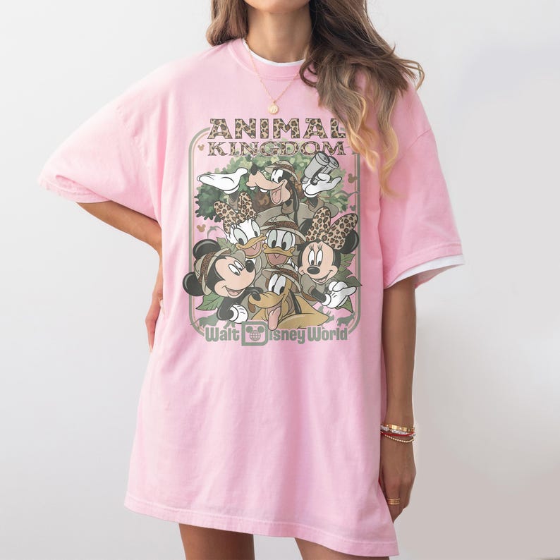 May include: Pink t-shirt featuring a graphic of Disney characters, including Mickey and Minnie Mouse, Goofy, Donald Duck, and Daisy Duck, with the text "Animal Kingdom" and "Walt Disney World". The shirt has white trim on the sleeves.