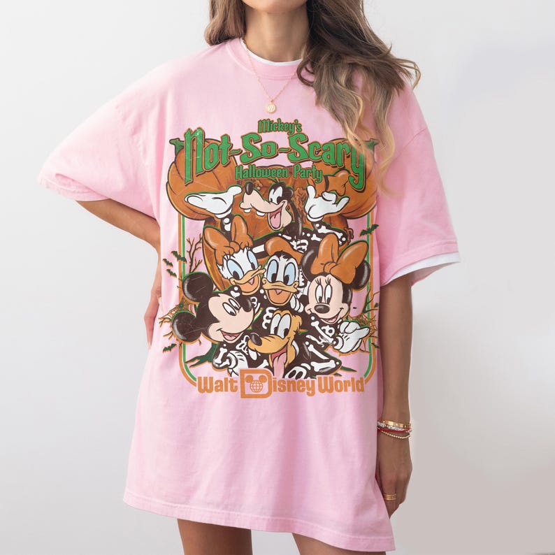 May include: A light pink t-shirt featuring a graphic print of Mickey Mouse, Minnie Mouse, Donald Duck, Daisy Duck, and Goofy wearing skeleton costumes. The text reads Mickey's Not-So-Scary Halloween Party and Walt Disney World in orange and green.