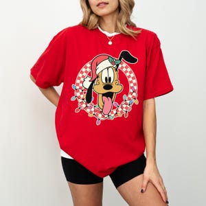 May include: Red t-shirt featuring a cartoon Pluto wearing a Santa hat and surrounded by a wreath of Christmas lights. The design includes a checkered pattern and Pluto's tongue sticking out. The shirt has a white collar.