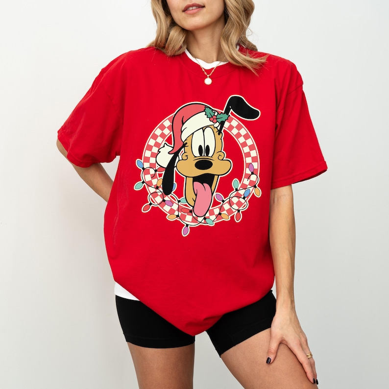 May include: Red t-shirt featuring a cartoon Pluto wearing a Santa hat and surrounded by a wreath of Christmas lights. The design includes a checkered pattern and Pluto's tongue sticking out. The shirt has a white collar.