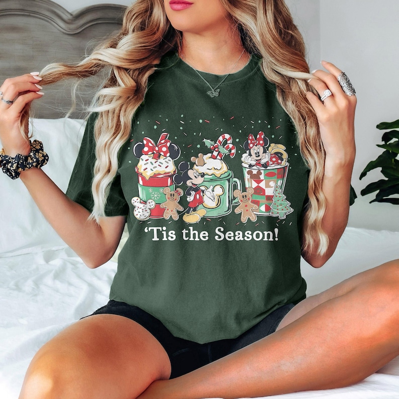 Puede incluir: Camiseta verde oscuro con un gr&aacute;fico festivo que presenta a Mickey y Minnie Mouse en tazas con crema batida y bastones de caramelo. Los hombres de jengibre y el texto "'Tis the Season!" completan el dise&ntilde;o navide&ntilde;o.