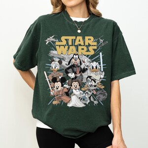May include: Dark green t-shirt featuring a graphic of Star Wars characters, including Mickey Mouse, Minnie Mouse, Donald Duck, and Goofy. The words "STAR WARS" are in gold lettering above the characters. The shirt has short sleeves and a crew neck.