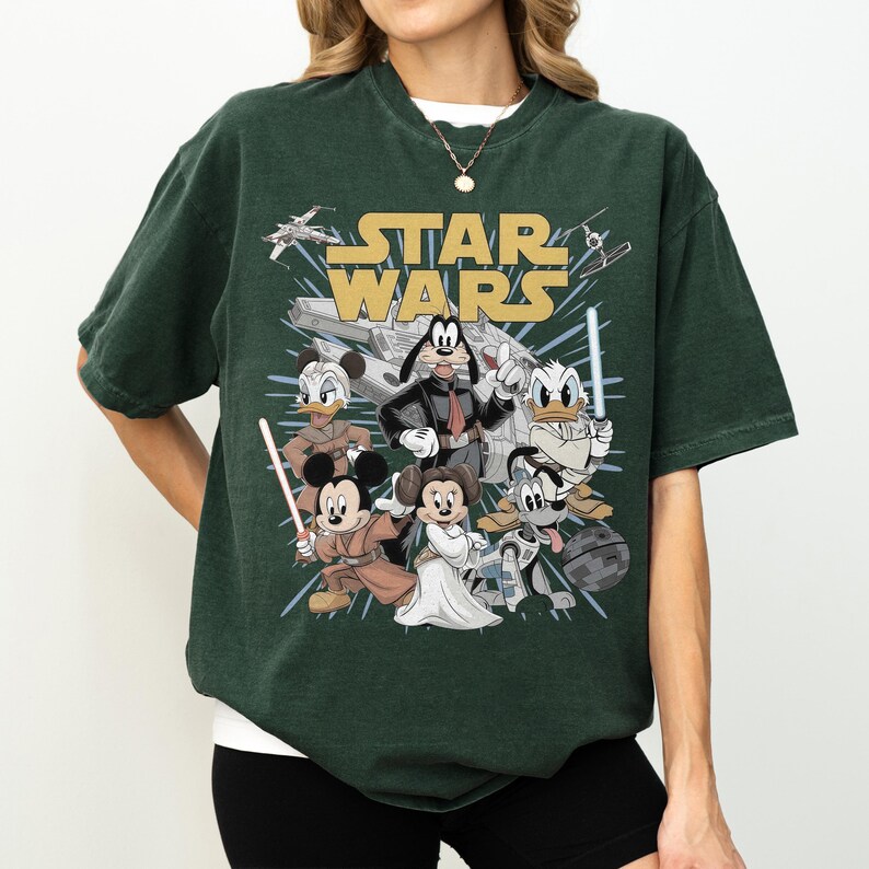 May include: Dark green t-shirt featuring a graphic of Star Wars characters, including Mickey Mouse, Minnie Mouse, Donald Duck, and Goofy. The words "STAR WARS" are in gold lettering above the characters. The shirt has short sleeves and a crew neck.