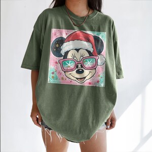 May include: Olive green t-shirt featuring a graphic of Mickey Mouse wearing a Santa hat and pink sunglasses with snowflake lenses. The graphic is set against a colorful, abstract background. The shirt has short sleeves and a relaxed fit.