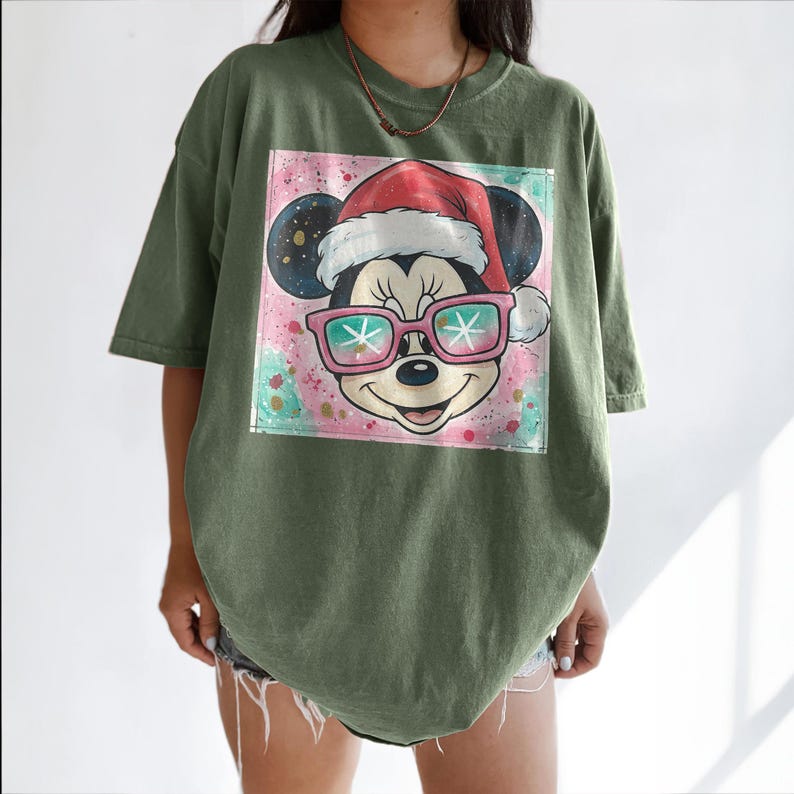 May include: Olive green t-shirt featuring a graphic of Mickey Mouse wearing a Santa hat and pink sunglasses with snowflake lenses. The graphic is set against a colorful, abstract background. The shirt has short sleeves and a relaxed fit.