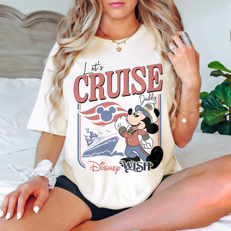 May include: Cream-colored t-shirt featuring the text "Let's Cruise Daddy" in red and white. The design includes a cartoon image of Mickey Mouse in a captain's uniform, a cruise ship, and the words "Disney Wish".
